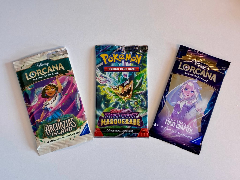 Three trading card game packs displayed on a white surface: 'Lorcana: Archazia's Island,' 'Pokémon: Twilight Masquerade,' and 'Lorcana: The First Chapter.'