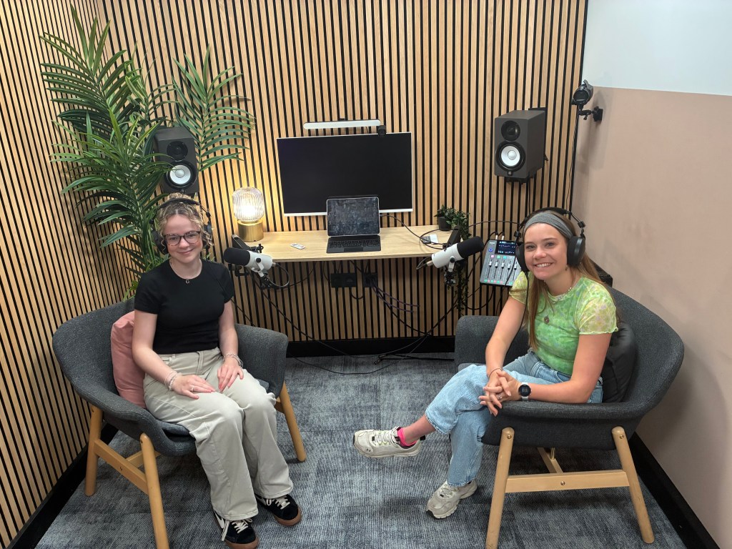 Two women sitting in a modern podcast studio, each wearing headphones and microphones, with a wooden wall background and plants in the corners.