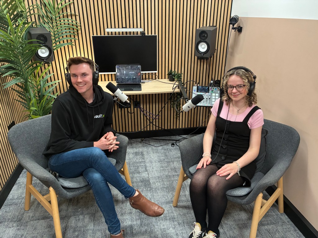 Two podcast hosts sitting in a modern recording studio, smiling and wearing headphones. They are seated in comfortable chairs with microphones in front of them, surrounded by audio equipment.