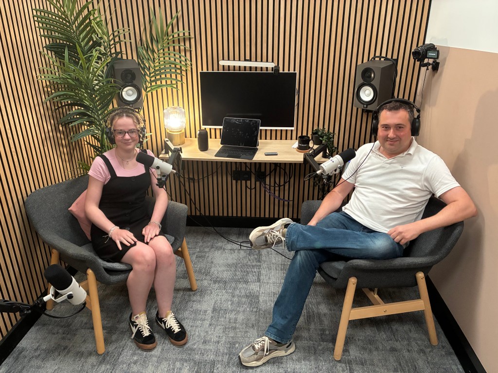 A woman and a man seated in a modern podcast studio, with microphones and a computer setup in the background. The studio features wooden paneling and decorative plants.