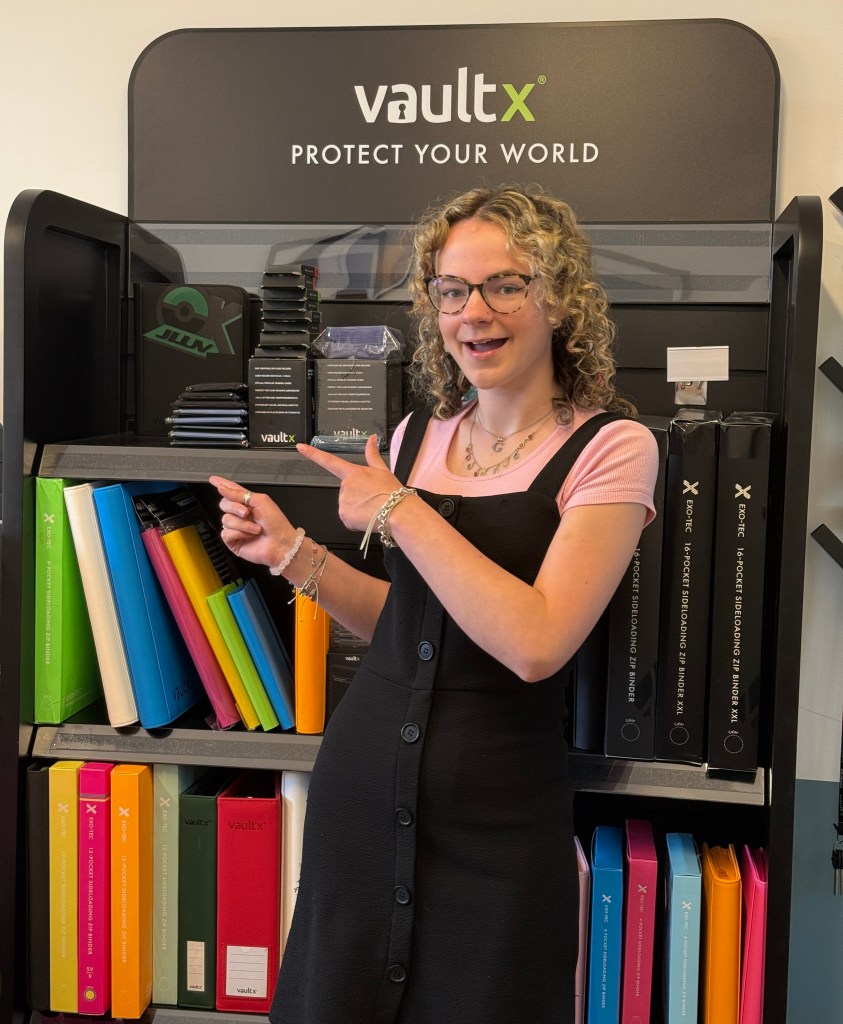 A young woman with curly hair and glasses is standing in front of a shelf displaying various colored binders and card protection products, pointing towards the products. The shelf is branded with 'vaultX' and the slogan 'PROTECT YOUR WORLD.'