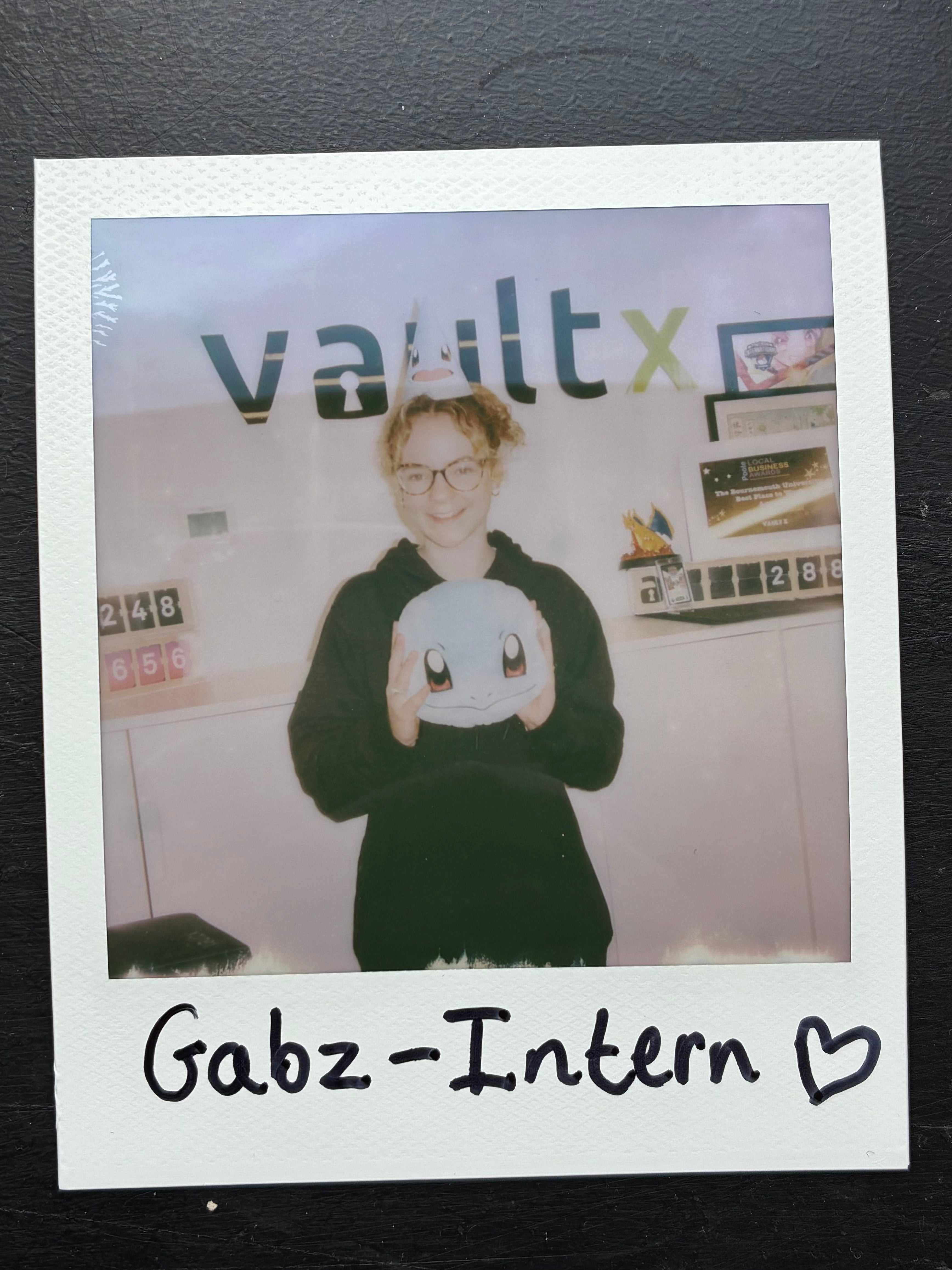 A polaroid photo of a smiling intern named Gabz holding a plush Pokémon character in front of the Vault X logo.