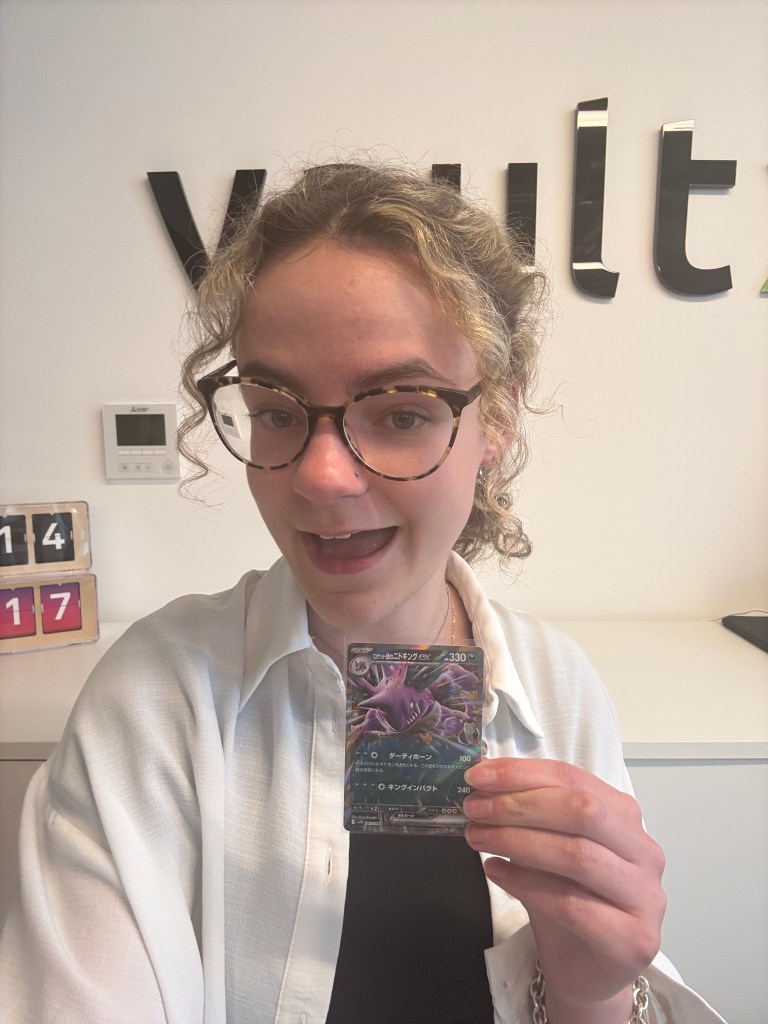 A person holding a Pokémon card featuring an illustration of a Pokémon, smiling in front of a wall with the word 'vault' in large letters.