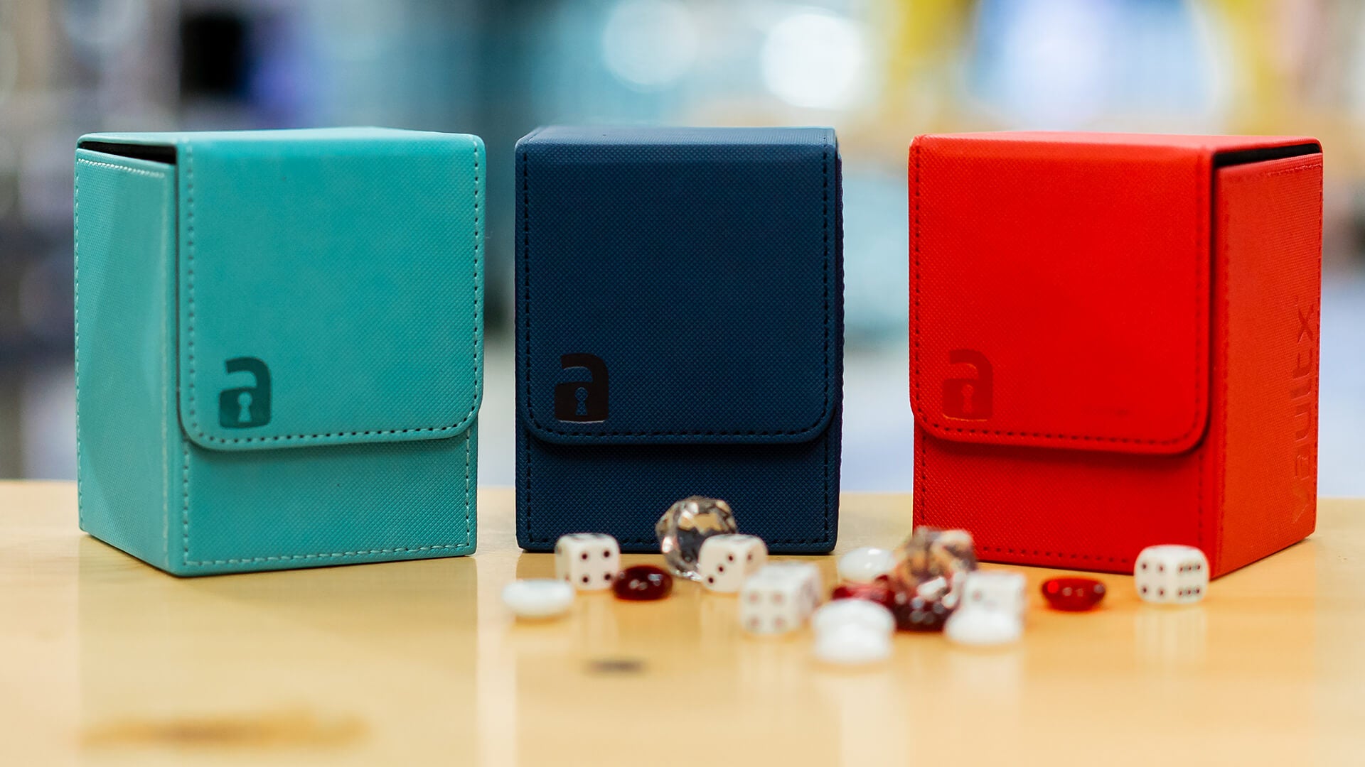 Three deck boxes in teal, navy, and red colors, positioned on a wooden surface with various game tokens and dice scattered around.