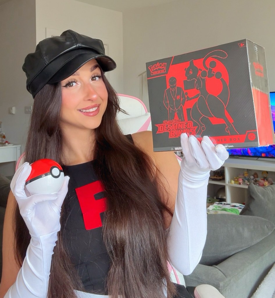 A content creator dressed in a Team Rocket costume holding a Pokémon box and a Poké Ball, smiling at the camera.