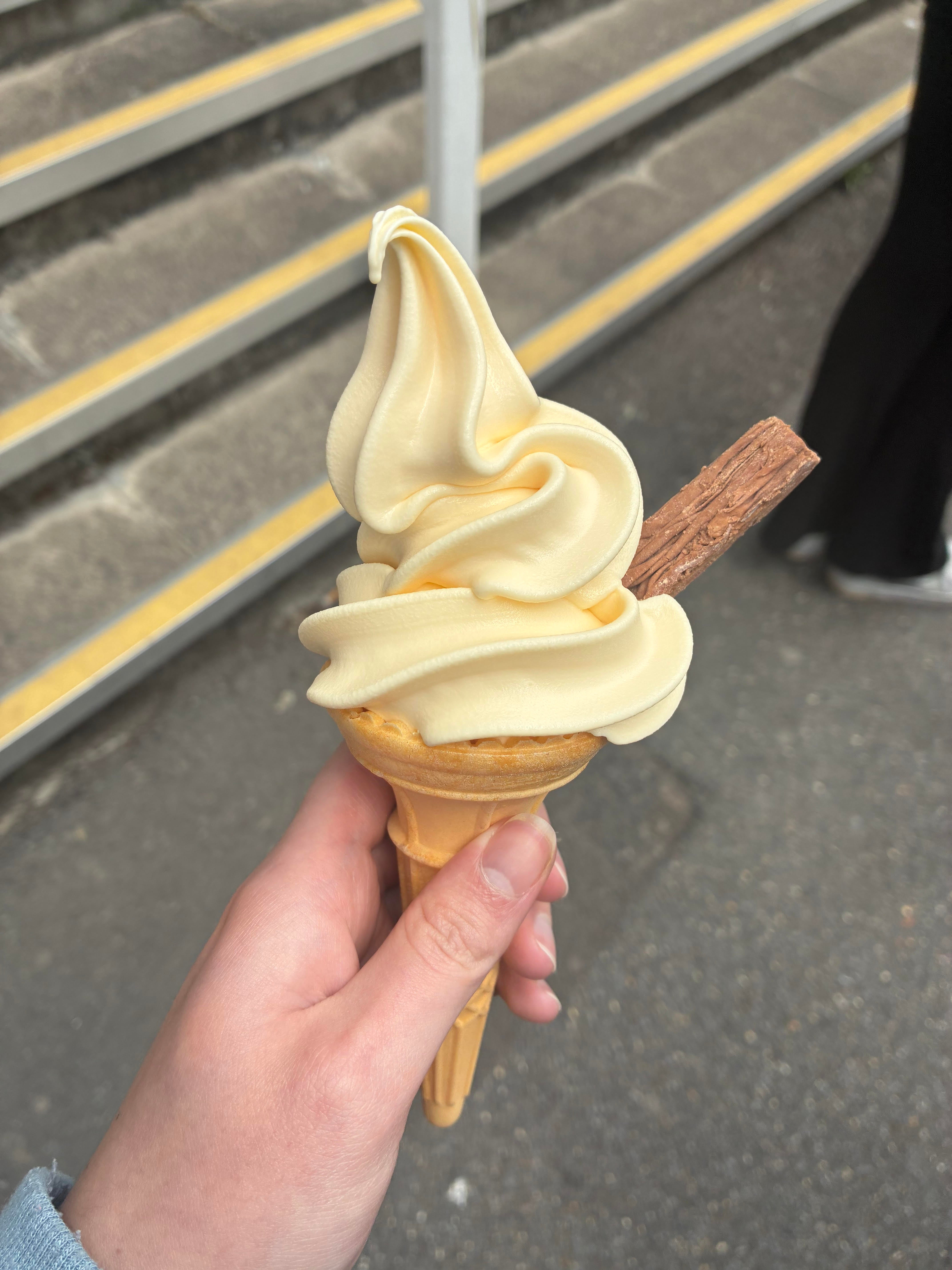 A hand holding a cone of soft serve ice cream topped with a chocolate stick, set against a background of stone steps.