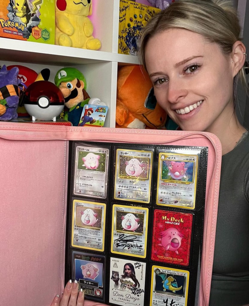 A person smiling while holding a binder displaying various Pokémon cards featuring multiple 'Chansey' and 'Blissey' cards, on a background with Pokémon plush toys and memorabilia.