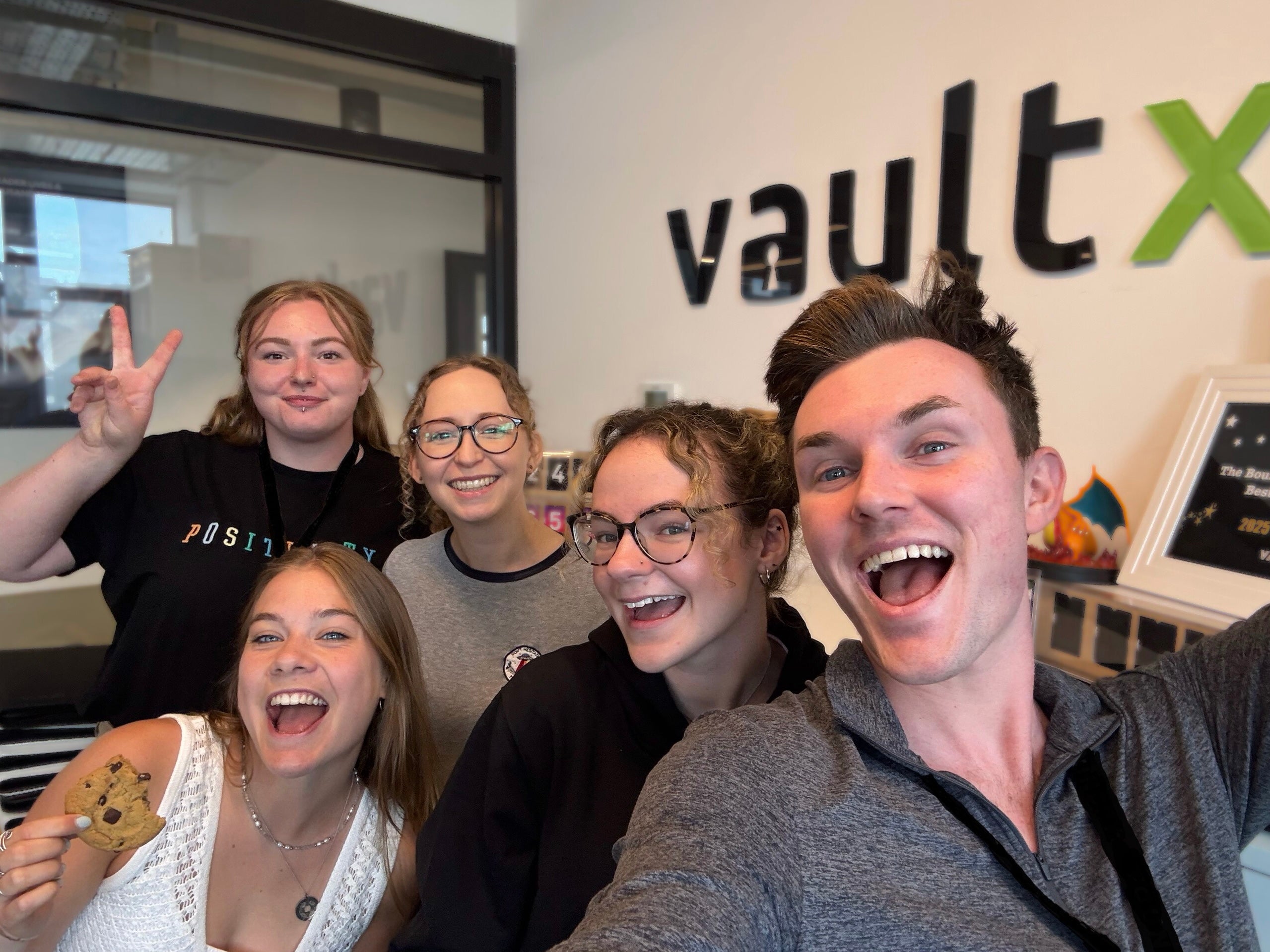 A group of five young adults smiling and posing for a selfie in an office environment, with the 'vaultX' logo visible in the background. One person is holding a cookie.