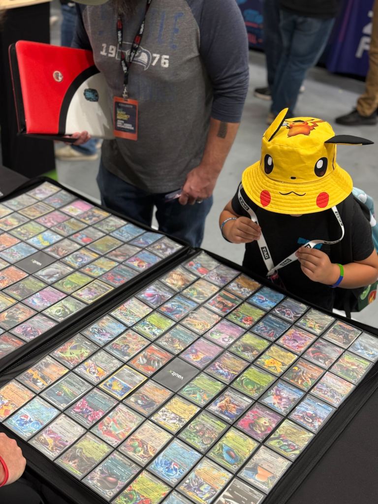 A child wearing a Pikachu bucket hat examines a display of collectible trading cards, while an adult observes nearby. The cards are organized in protective sleeves on a table.