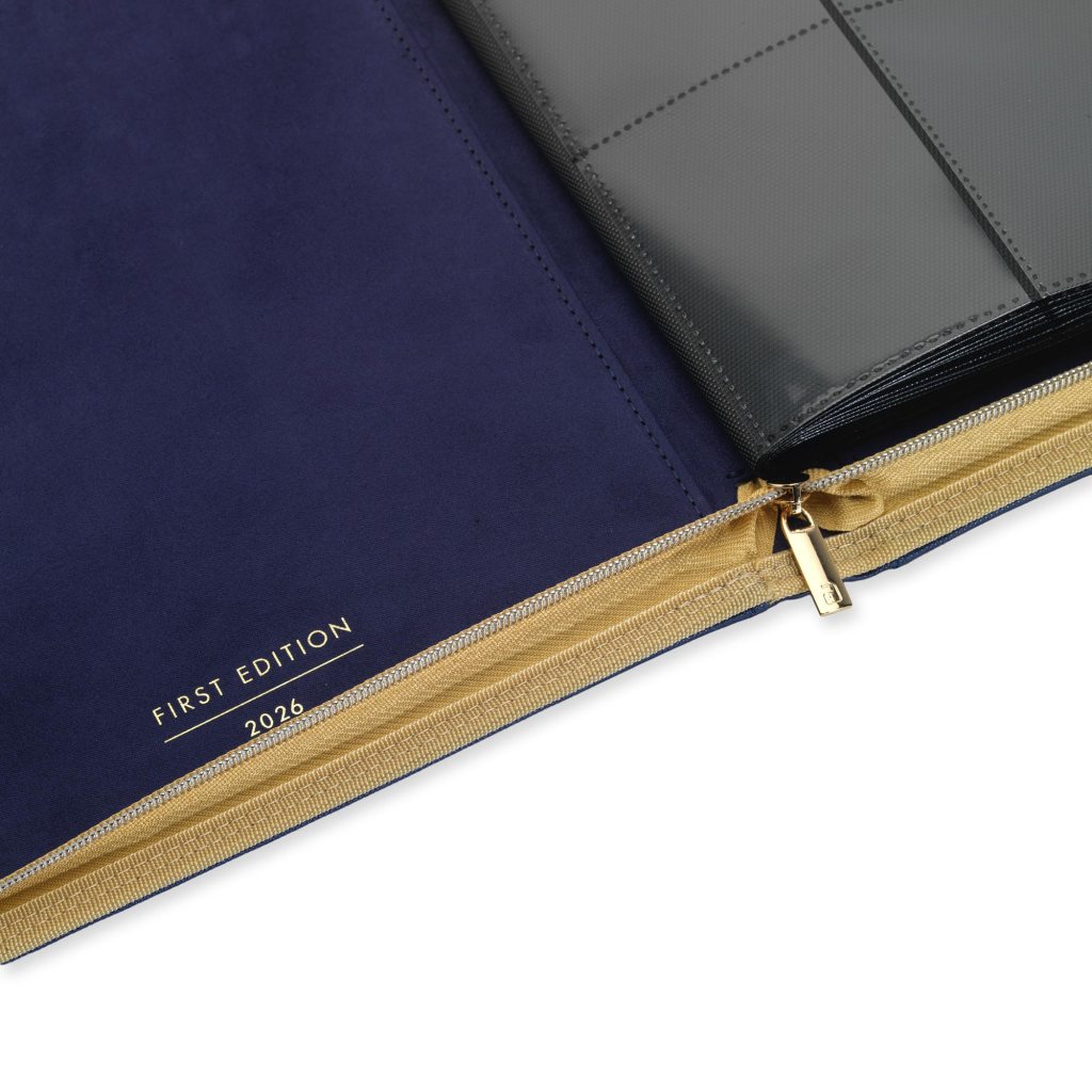 Close-up of a navy blue folder with 'FIRST EDITION' and '2026' embossed in gold, featuring a gold zipper and a clear plastic insert.