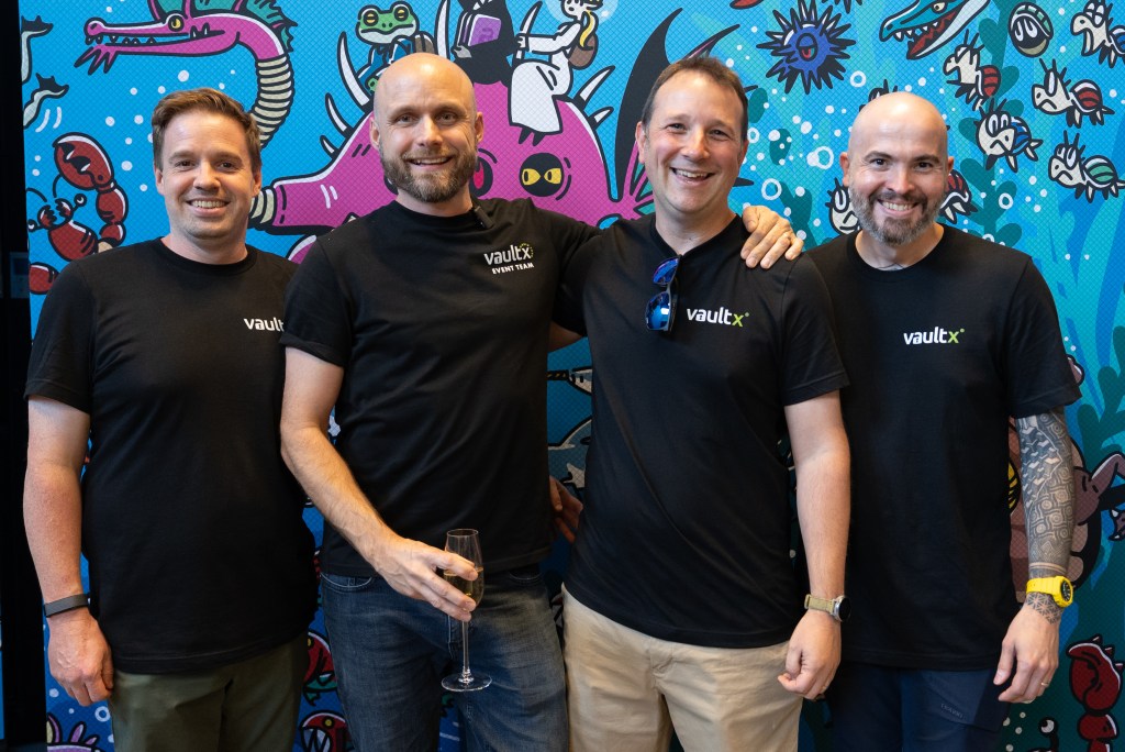 Four men in black t-shirts with the logo 'vaultx' stand together in front of a colorful cartoon backdrop featuring underwater themes.