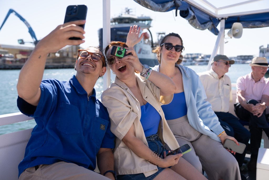 A group of friends taking a selfie on a boat, with a waterfront and industrial backdrop. They are smiling and enjoying the moment, with one person holding a vintage camera.