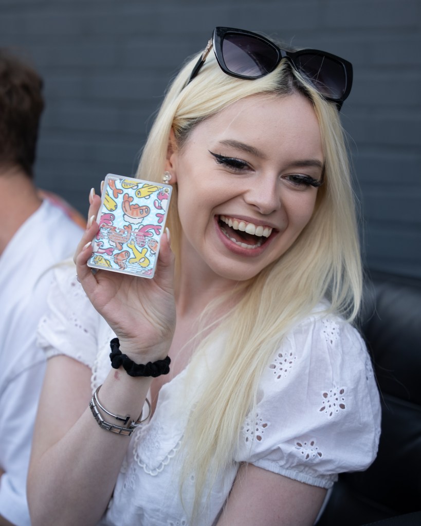 A smiling young woman with long blonde hair holds up a colorful decorated box while wearing sunglasses and a white dress.