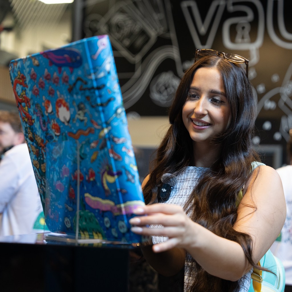 A woman smiles while examining a colorful illustrated book featuring underwater scenes, inside a lively browsing space.